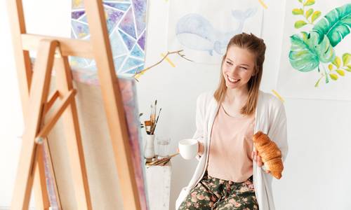 Frau sitzt lachend vor Staffelei und hält eine Tasse Kaffee in der Hand